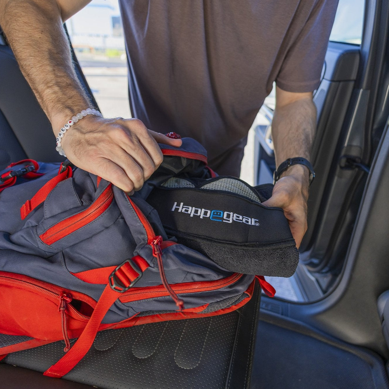Close-up of the black Happeseat headrest strap with logo being placed on a car seat beside a gray and red backpack